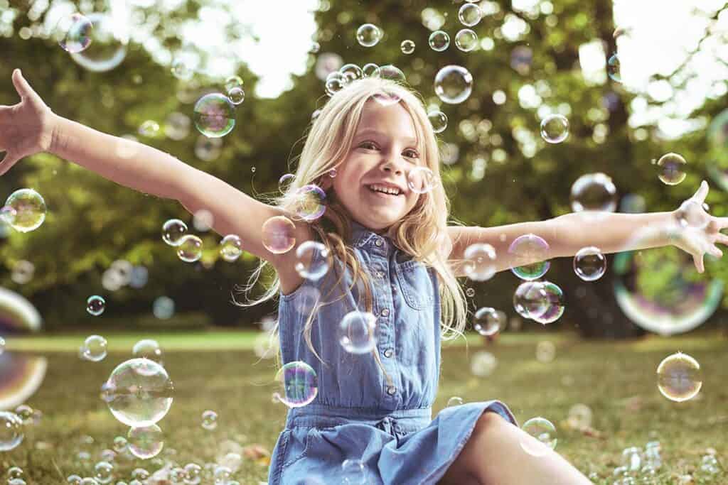 Child smiling with their arms spread wide, as large soap bubbles float all around them.