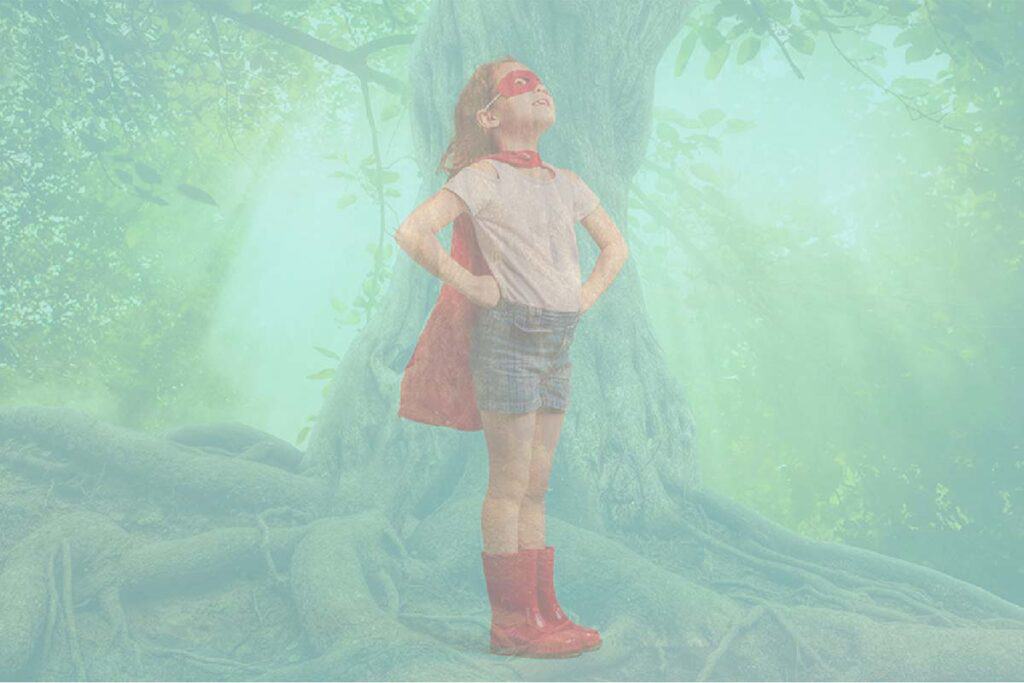 Child wearing a cape, eye mask, and rain boots, standing with their hands on their hips, in front of a large old tree with very large roots.