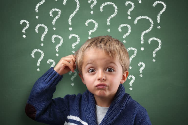Confused child scratching their head, while standing in front of a chalkboard filled with question marks.