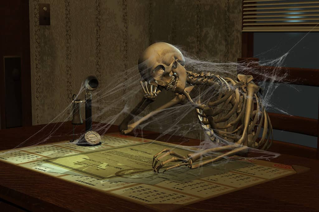 A cobweb-filled skeleton sits at a desk beside an old-fashioned phone and stares at a desktop calendar.