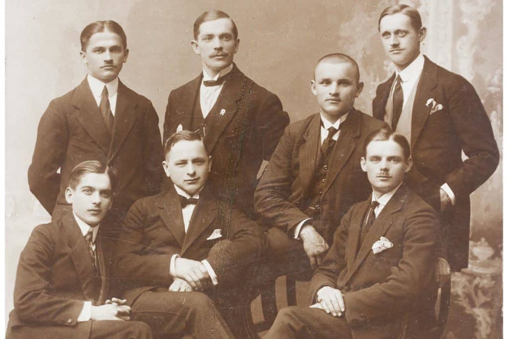 Early photo of seven young white men in suits, posing for the camera.