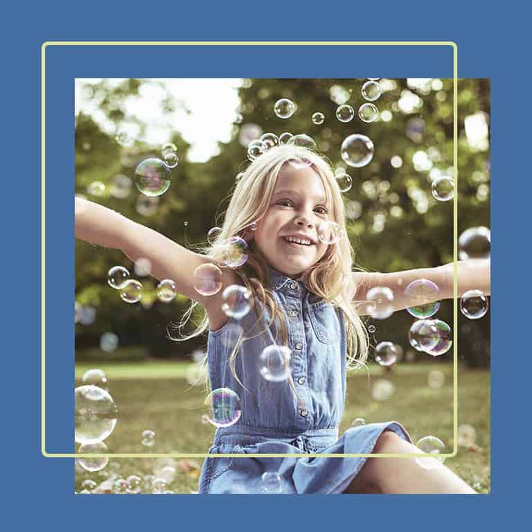 Feel like you belong within this fluid bubble. A happy child sits in a meadow, her arms wide apart, surrounded by bubbles.